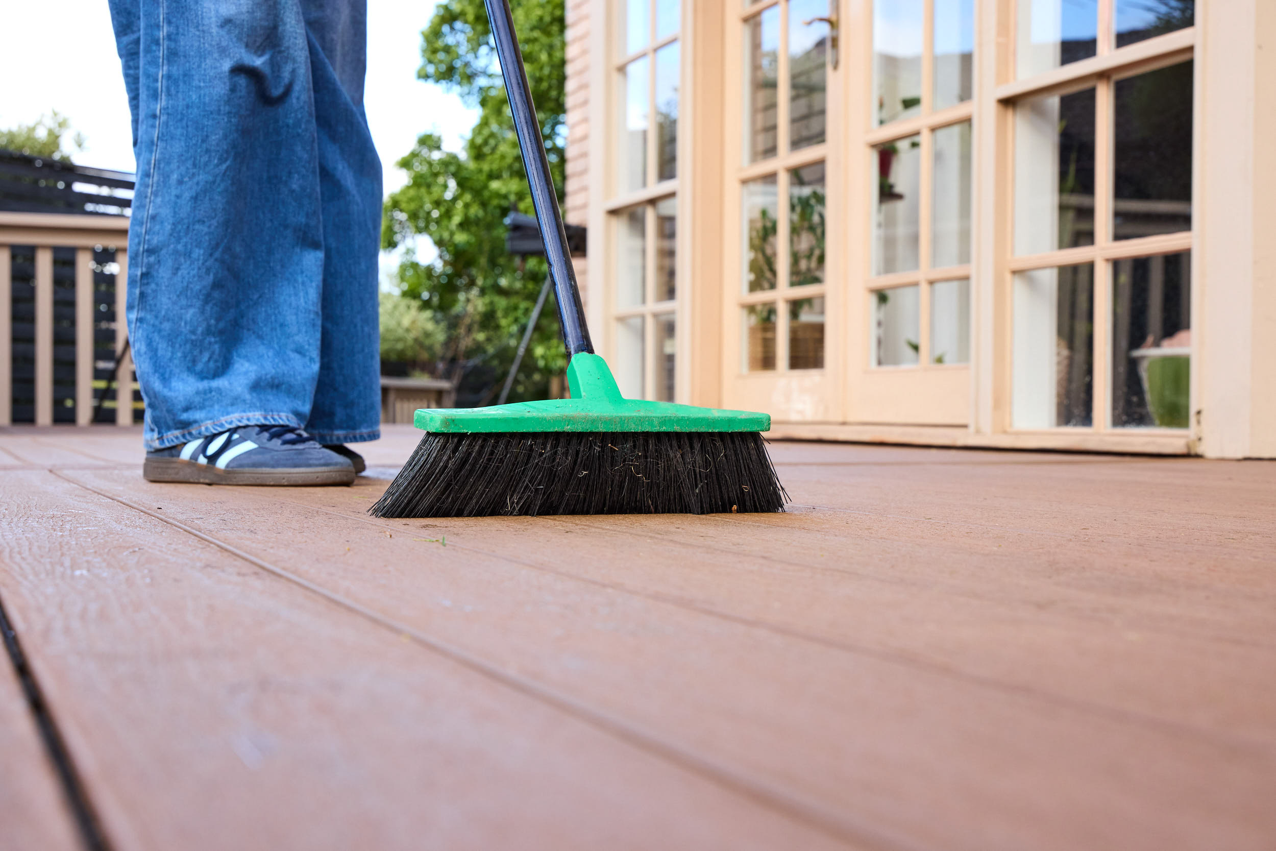 A broom with a green handle and black bristles being pushed on a brown deck