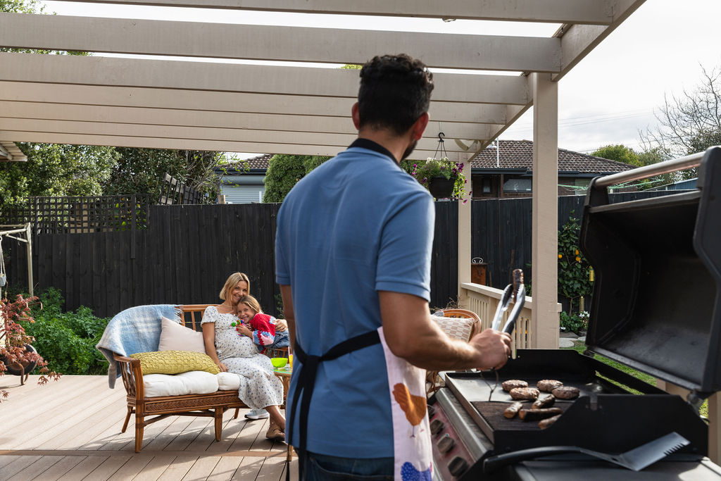 A man cooking at a barbecue looking at a pregnant woman and child relaxing on an outdoor couch